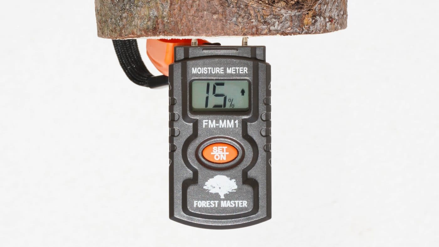 Black Forest Master Moisture meter with a white logo at the bottom and an orange button in the centre, pinned to a log with LCD screen displaying fifteen per cent.